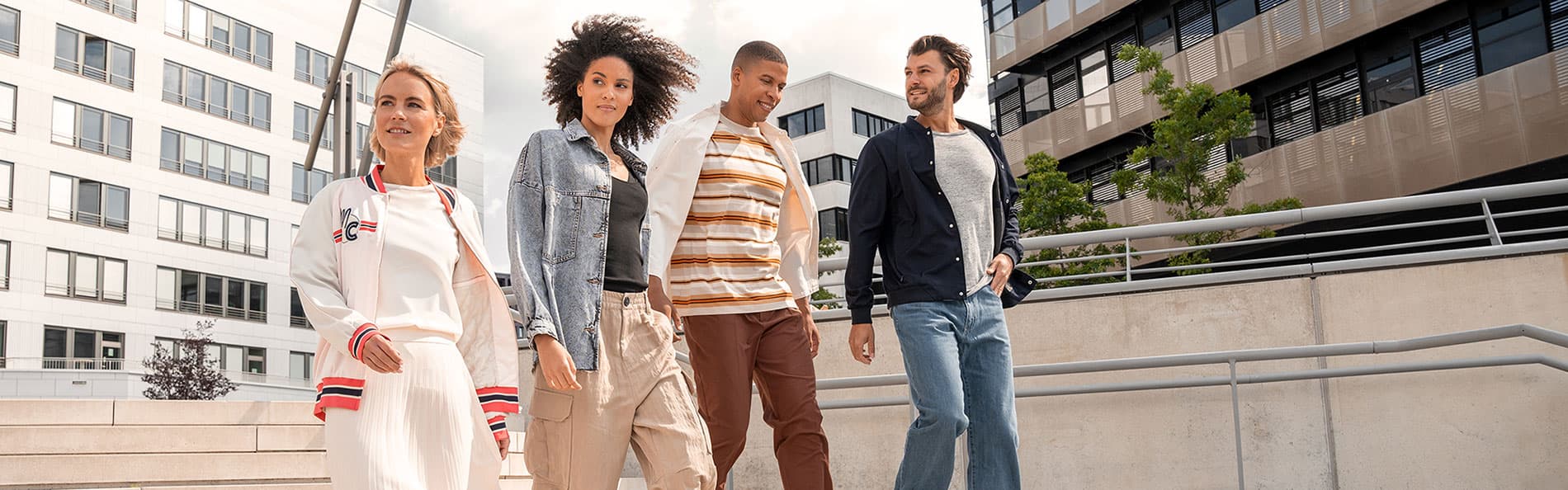 Vier Personen gehen fröhlich eine Treppe hinunter in einer städtischen Umgebung mit modernen Gebäuden im Hintergrund. Sie sind leger gekleidet und lächeln.