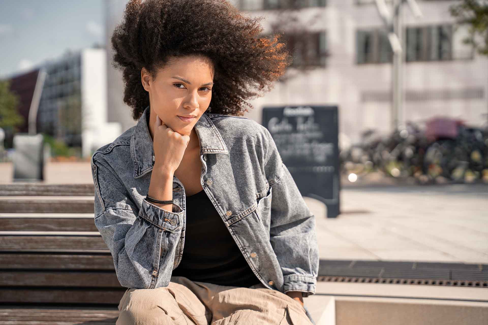 Frau in Jeansjacke, die auf einer Außentreppe sitzt, die Hand unter dem Kinn, mit Gebäuden und Fahrrädern im Hintergrund.