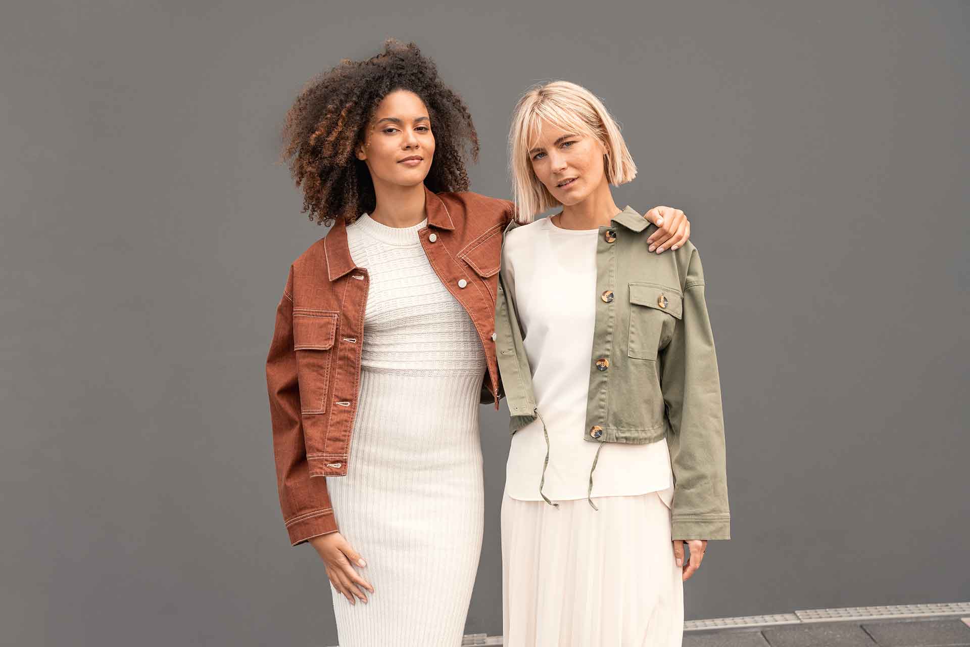 Zwei Frauen stehen dicht beieinander und lächeln. Die eine trägt eine braune Jacke über einem weißen Kleid, die andere eine olivgrüne Jacke über einem weißen Outfit. Der Hintergrund ist grau.