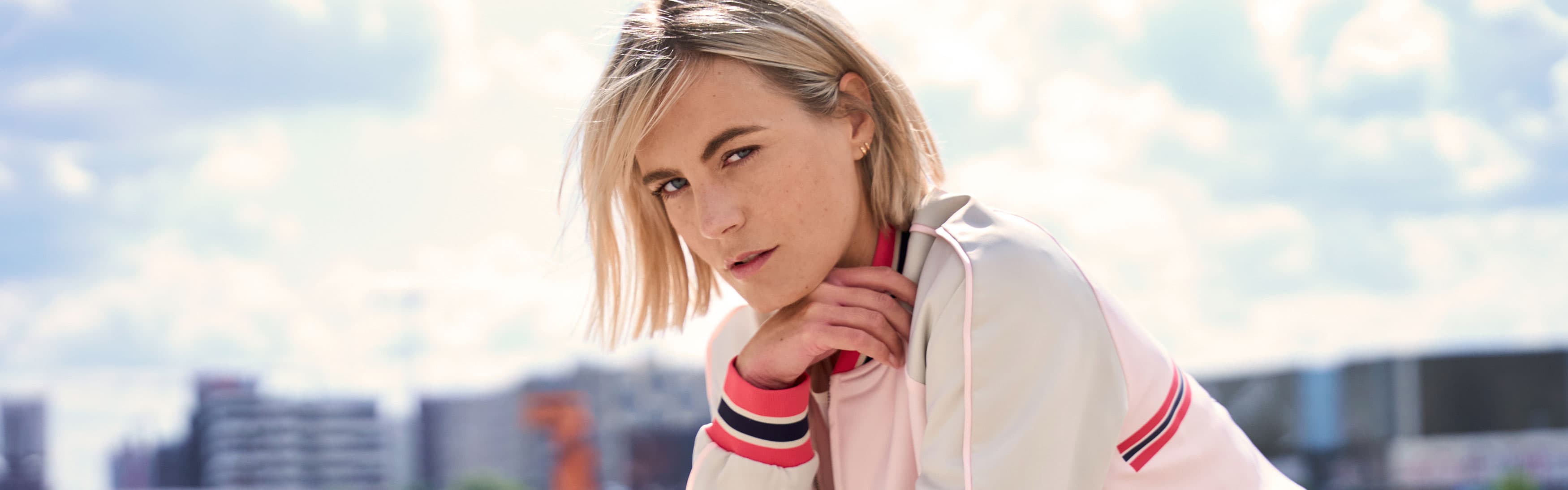Eine Frau mit blonden Haaren in einer weißen Jacke blickt aufmerksam in die Kamera, im Hintergrund ist eine Stadtlandschaft zu sehen.