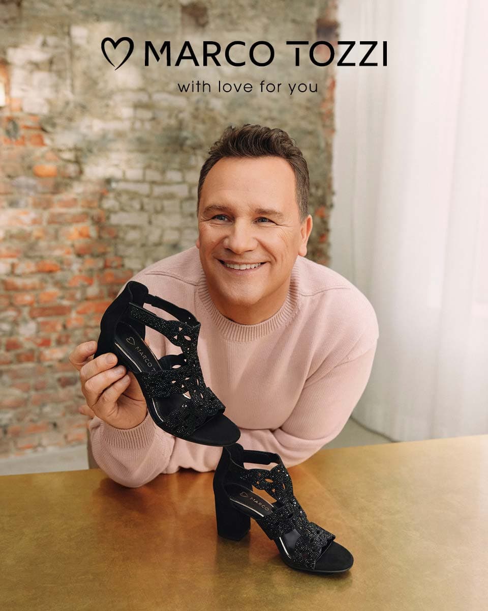 Ein lächelnder Mann (Guido Maria Kretschmer) in einem rosa Pullover, der einen schwarzen Riemchen-High-Heel von MARCO TOZZI auf einem goldenen Tisch hält, vor dem Hintergrund einer Backsteinmauer.