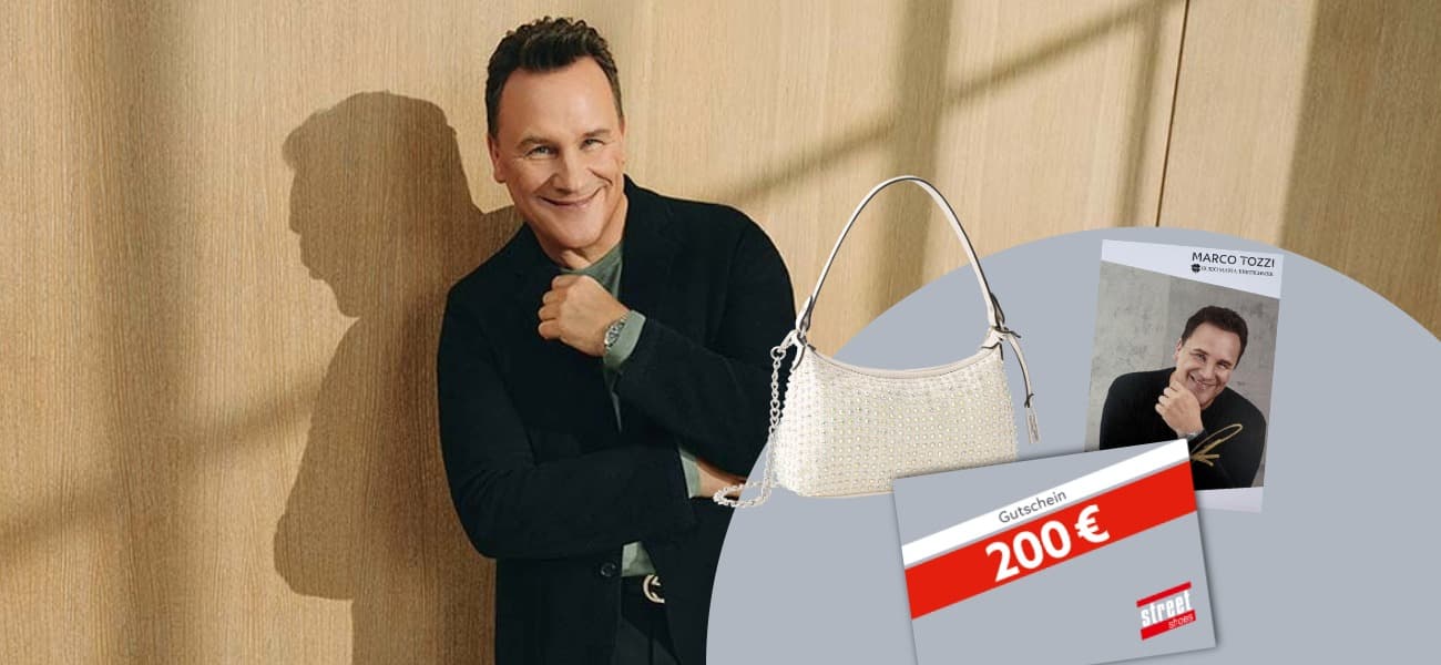 Ein lächelnder Mann (Guido Maria Kretschmer) in dunkler Kleidung, eine Handtasche, ein signiertes Foto und ein 200-Euro-Gutschein vor einem holzfarbenen und grauen Hintergrund.