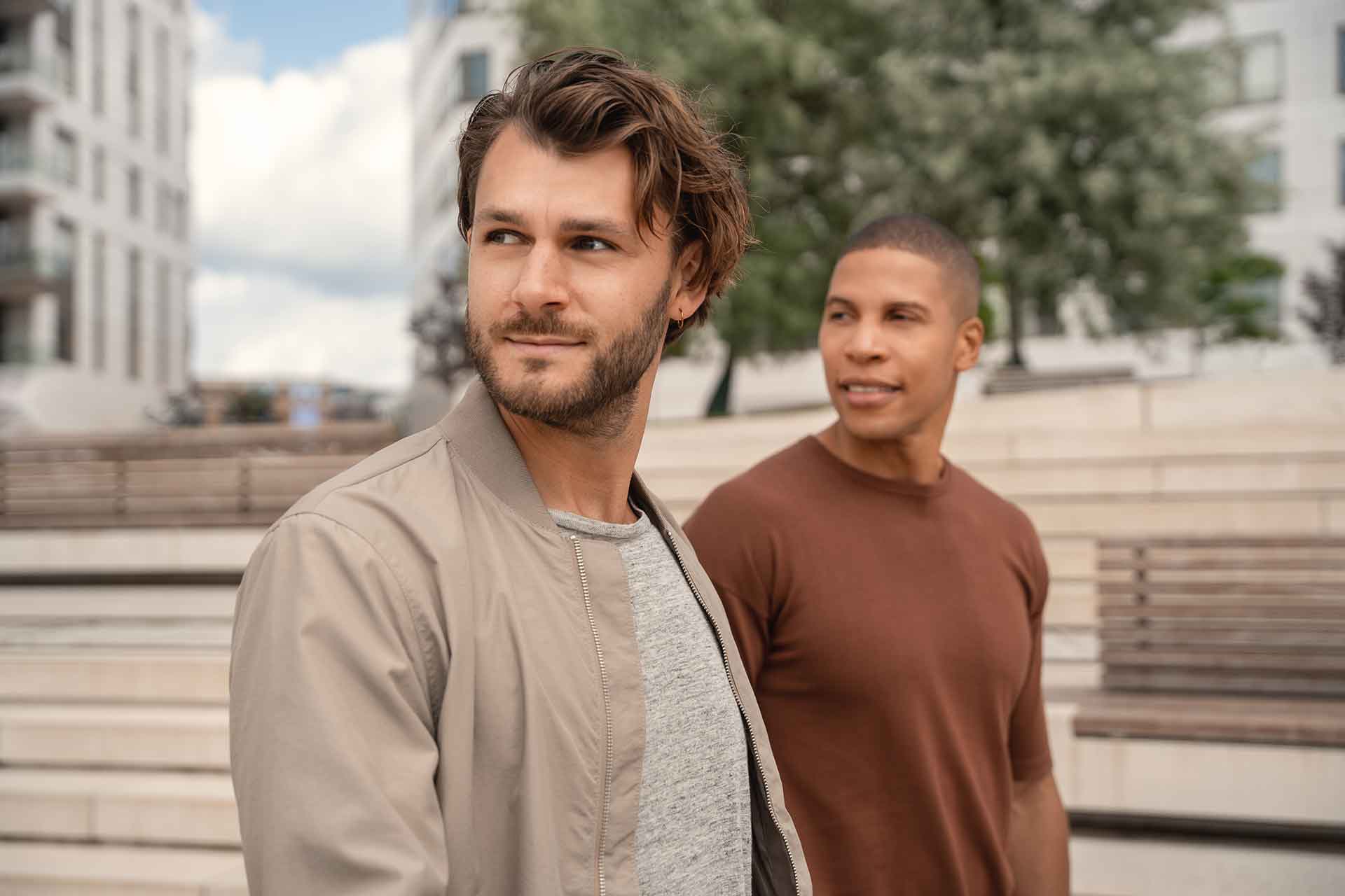 Zwei Männer stehen im Freien, im Hintergrund moderne Gebäude und Bäume. Beide schauen zur Seite.