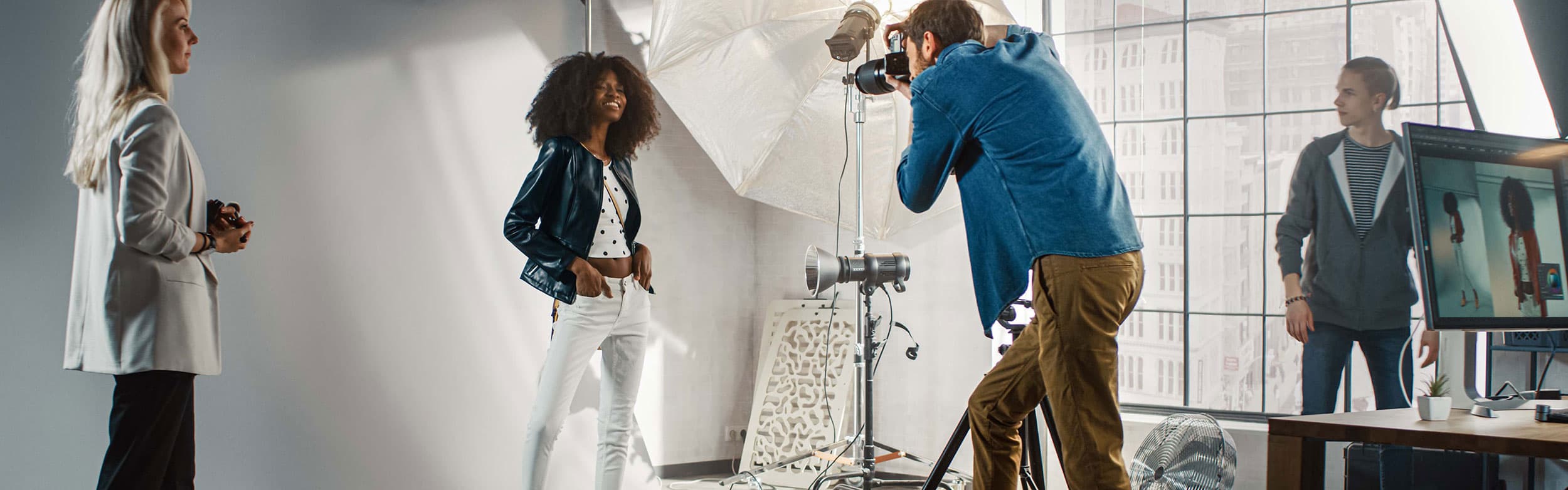 Model posiert in einem Studio, während ein Fotograf Bilder aufnimmt, zwei Assistenten, große Fenster und Beleuchtungsausrüstung im Hintergrund.