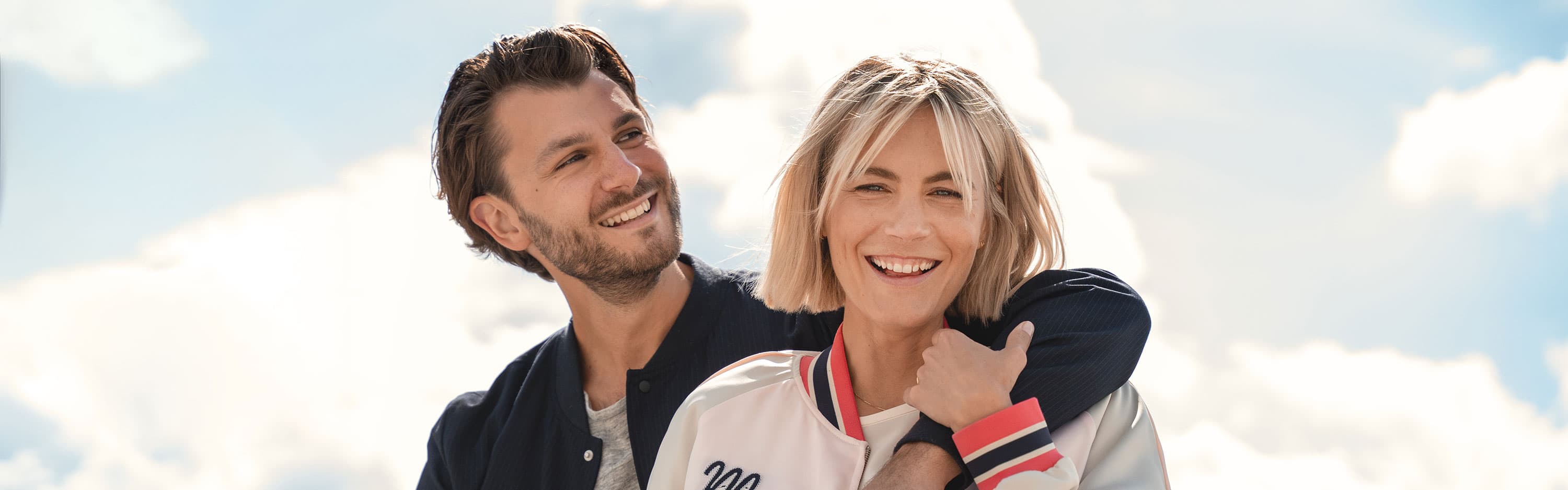 Ein Mann und eine Frau lächeln im Freien, der Mann hat seinen Arm um ihre Schultern gelegt, im Hintergrund sind blauer Himmel und Wolken zu sehen.